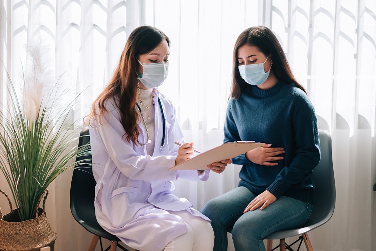 Asian doctor visit and examines on young adult woman at hospital with stomach ache. The doctor checking up and consulting for health care, wearing a mask to protect covid-19 pandemic.
